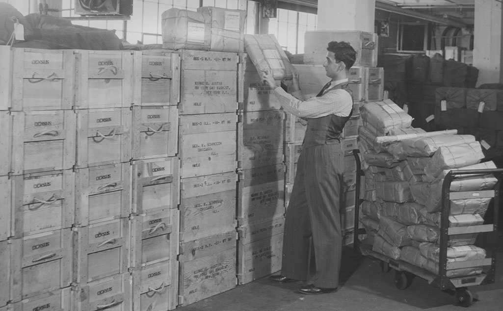a census bureau employee stands in front of multiple stacks of crates and bags, all filled with paper forms to be mailed all over