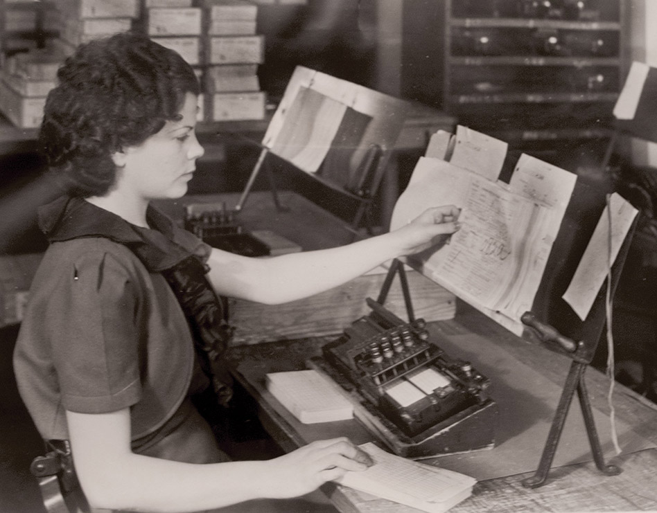 Photograph of a woman holding a sheaf of forms in one hand and a stack of paper cards in the others, while a card punch machine sits on the desk before her.