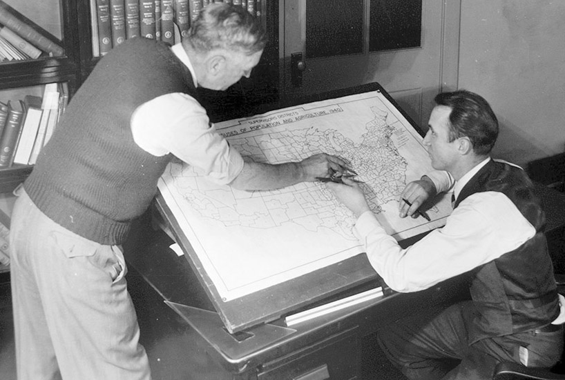 photo of two white men in dress shirts and vests, one with grey hair and the other brown, leaning over a drafting table that supports a map of the continental United States